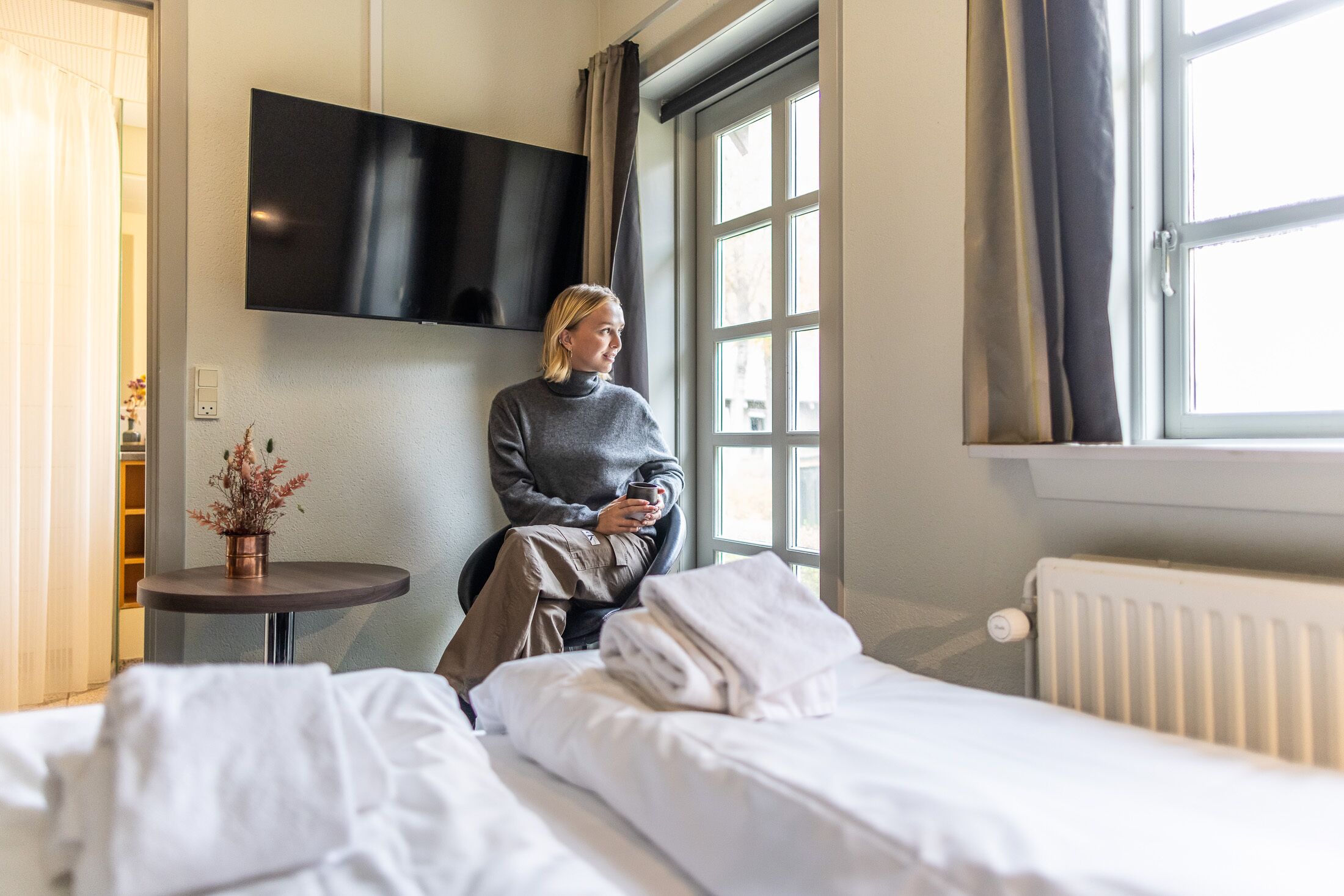 Habitación de hotel con cama, una mesita con una planta, televisor montado en la pared y una ventana con cortinas.