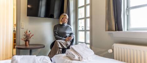 A hotel room with a bed, a small table with a plant, a TV mounted on the wall, and a window with curtains.