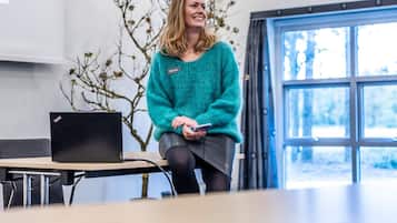 A person sitting in a room with a laptop on a table, a window with curtains, and a plant in the background.