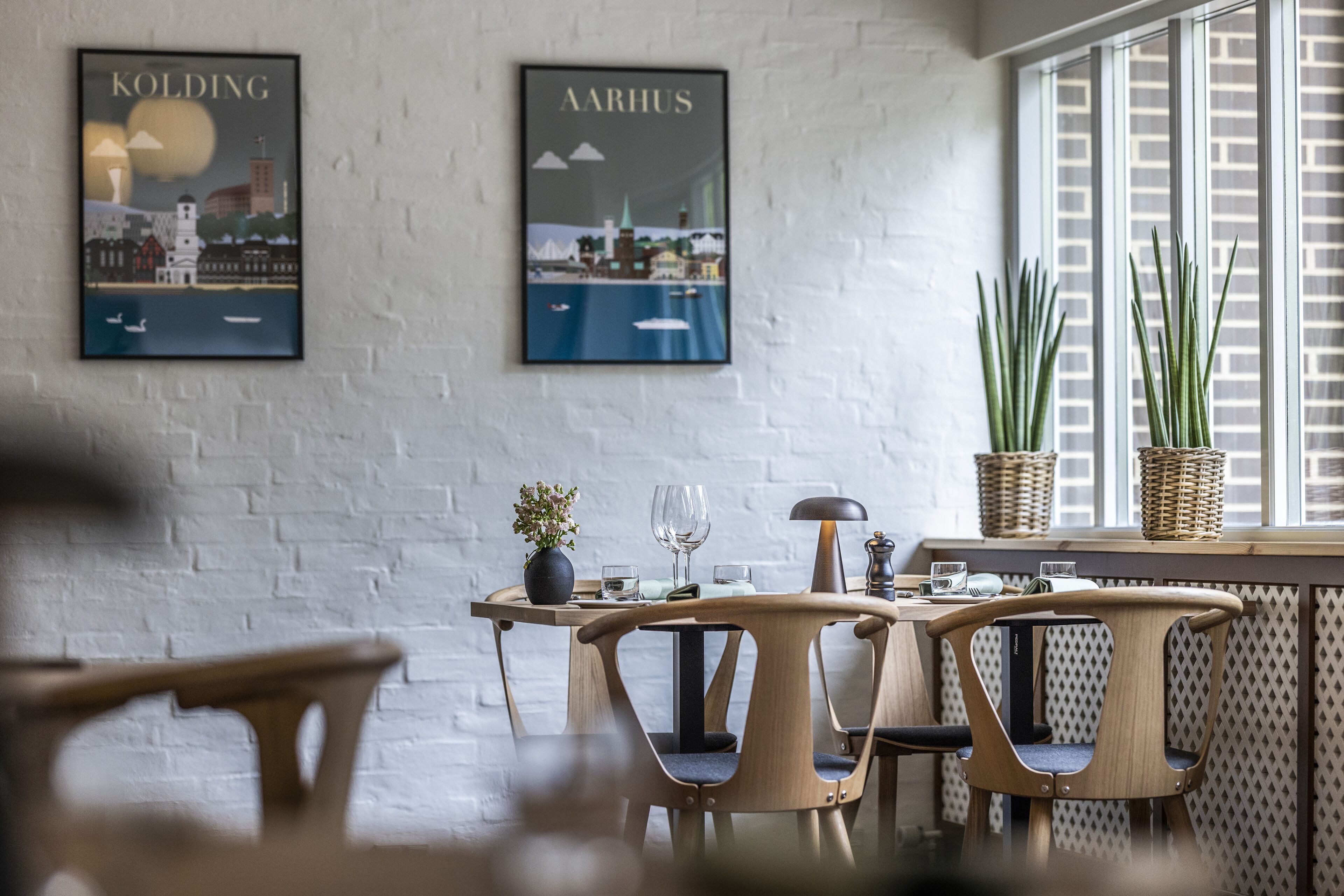 A dining area with a white brick wall, two framed cityscape posters, a table with chairs, a potted plant, and a window with blinds.