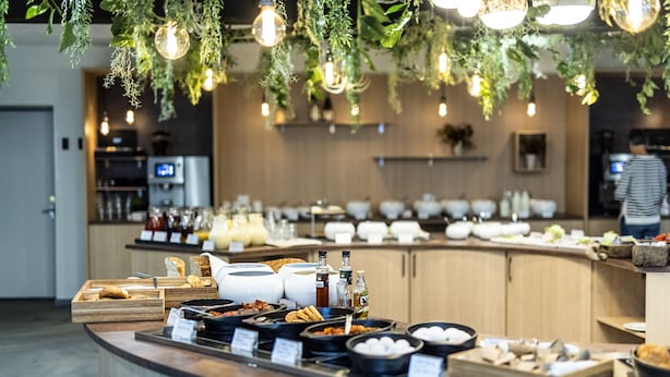 A buffet setup with various food items and beverages, a counter with a wooden finish, and hanging plants with lights.
