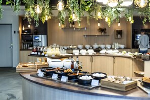 A buffet setup with various food items and beverages, a counter with a wooden finish, and hanging plants with lights. - The Lodge Billund (Billund)