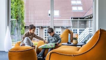 Dos niños jugando a un juego de mesa en una habitación moderna con ventanas amplias, muebles anaranjados y vistas a un edificio con tejados rojos.