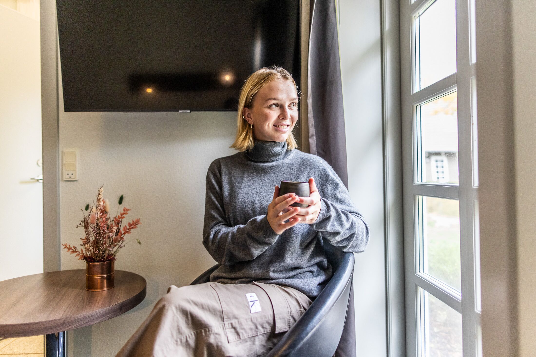 Una persona sentada junto a una ventana con vistas al exterior, sosteniendo una taza.