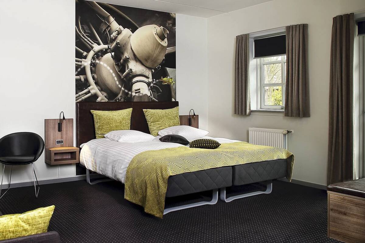 Moderni hotellihuone, jossa on suuri sänky, erikoisella designilla varustettu pääty, yöpöytä, tuoli ja ikkuna, jossa on verhot.