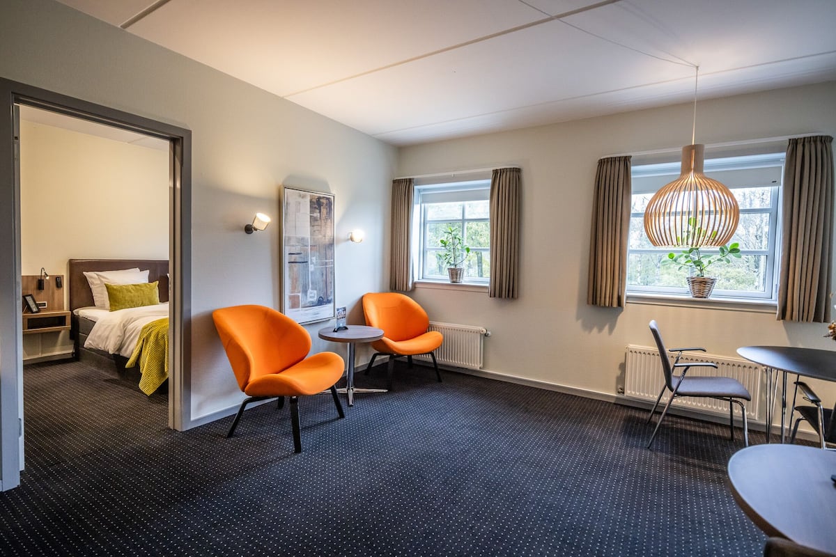 Hotellihuone, jossa on sänky, kaksi oranssia tuolia, pieni pöytä, ikkuna verhoilla ja riippuvalaisin.