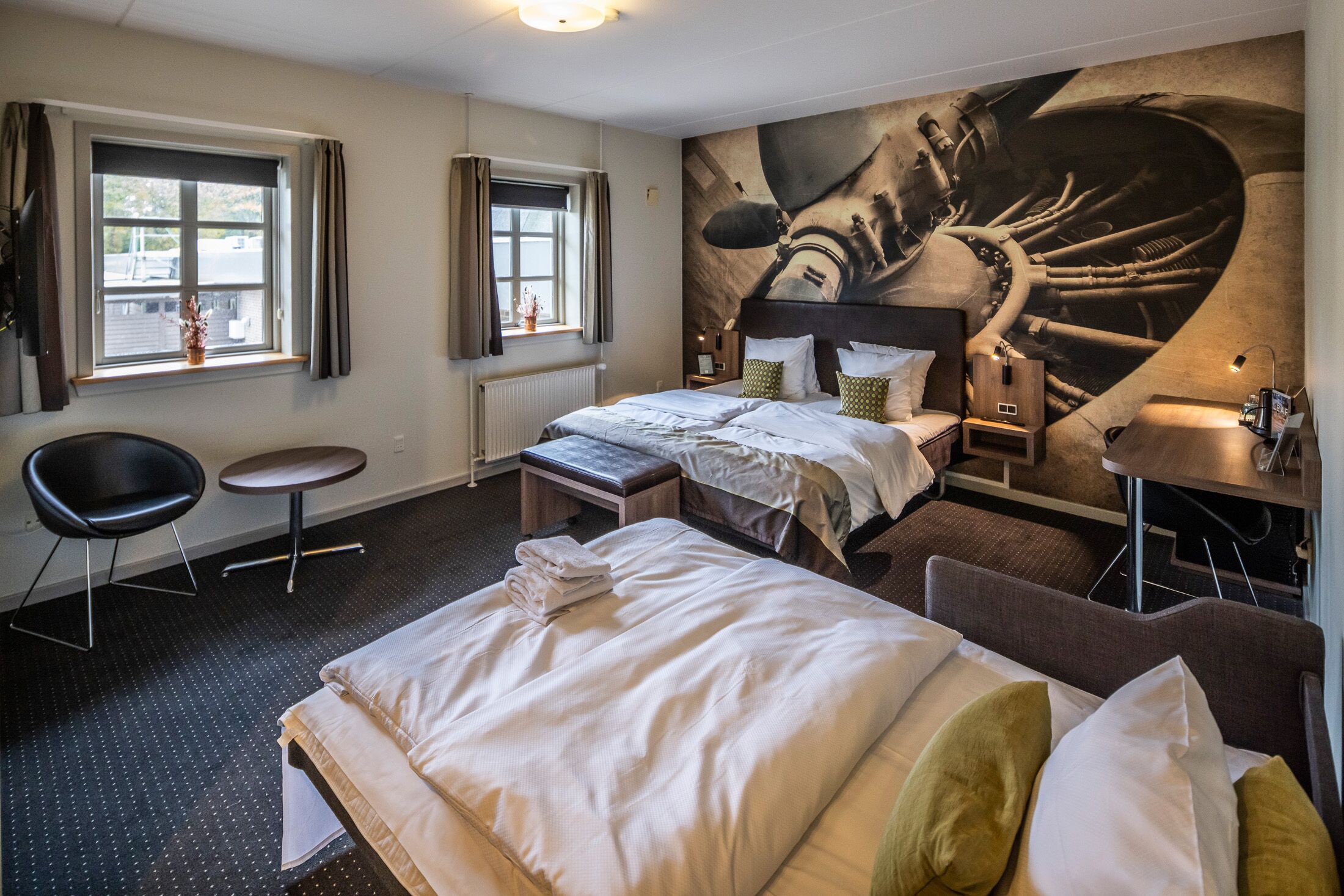 Een hotelkamer met een groot bed, twee nachtkastjes, een stoel, een klein tafeltje en een grote muurschildering.