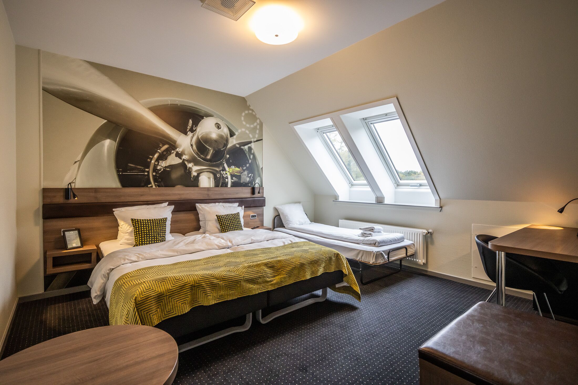 A hotel room with a large bed, a smaller bed, a desk, a chair, a window, and a ceiling light.