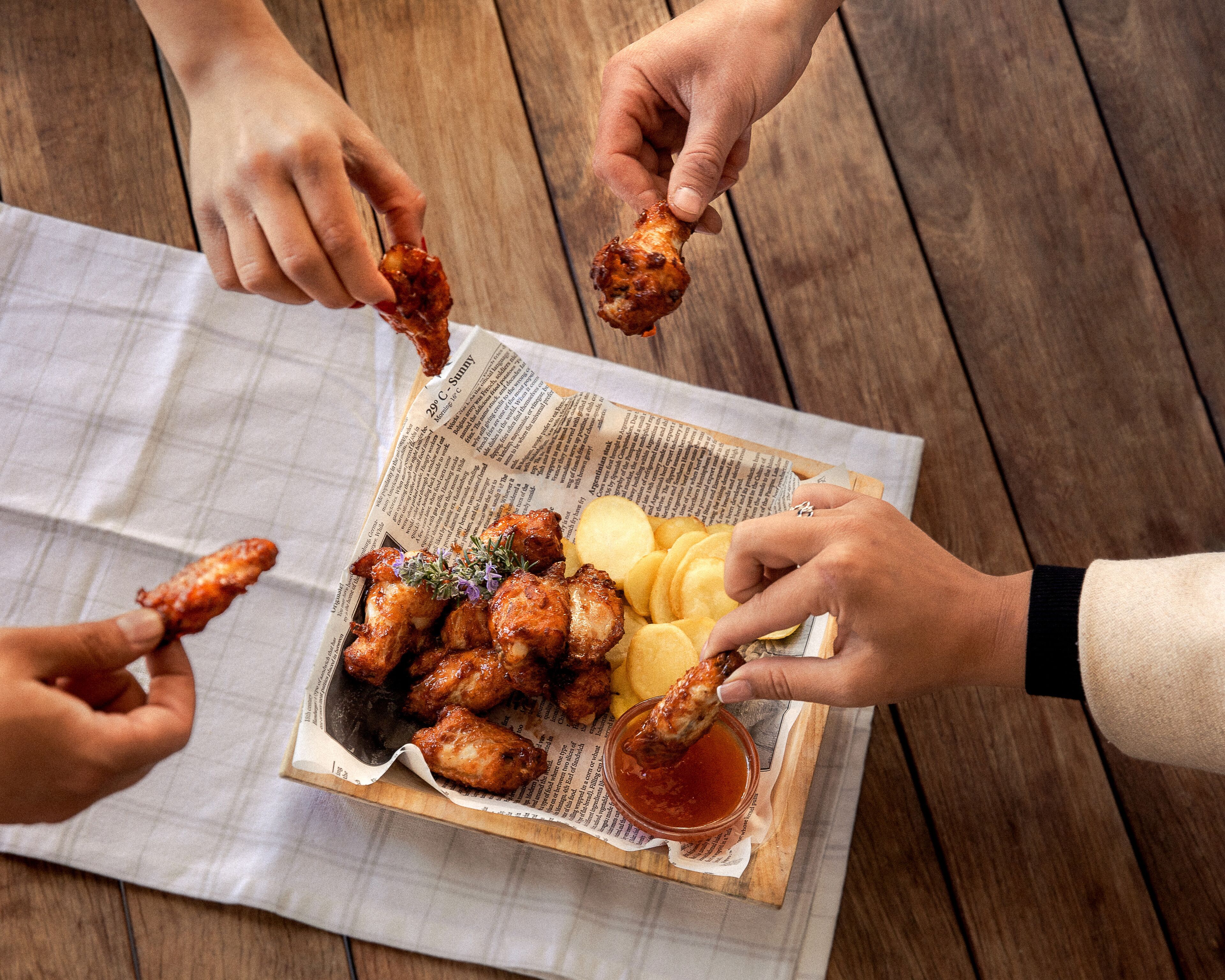 A wooden table with a newspaper-lined tray containing chicken wings, potato wedges, and a dipping sauce.