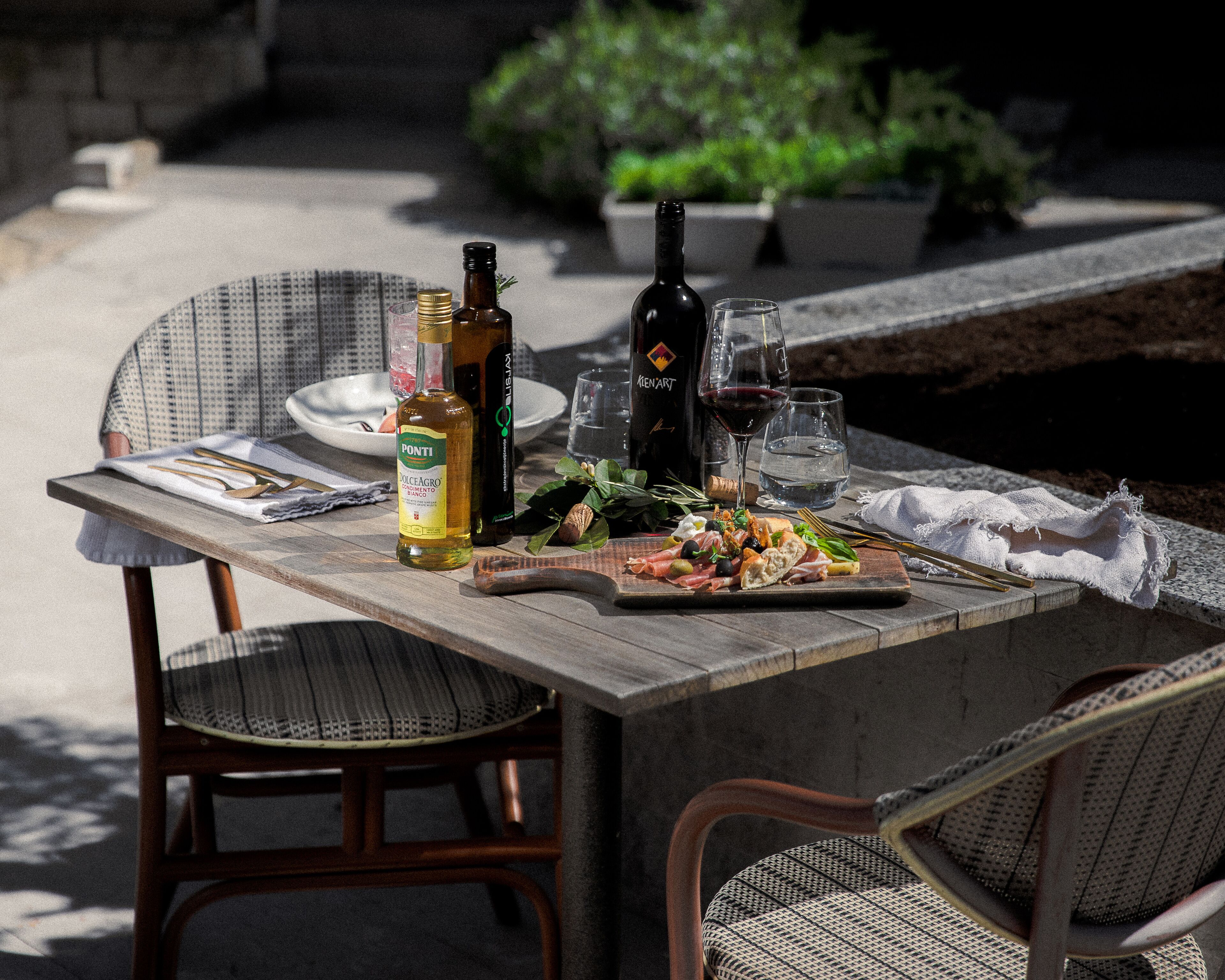 Een buitentafel met een fles wijn, glazen en een bord eten.