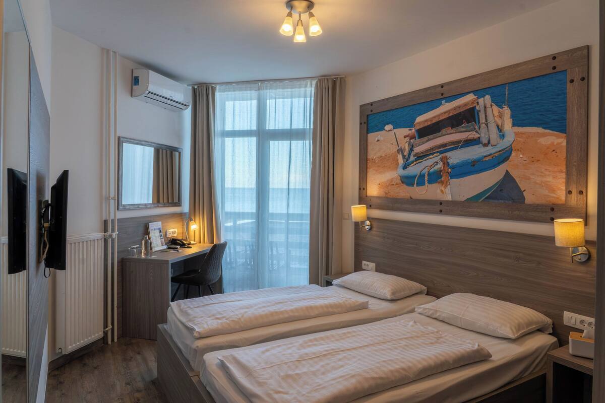 Une chambre d’hôtel avec deux lits, un bureau, une chaise, un appareil de climatisation, un tableau représentant un bateau et une fenêtre avec des rideaux.