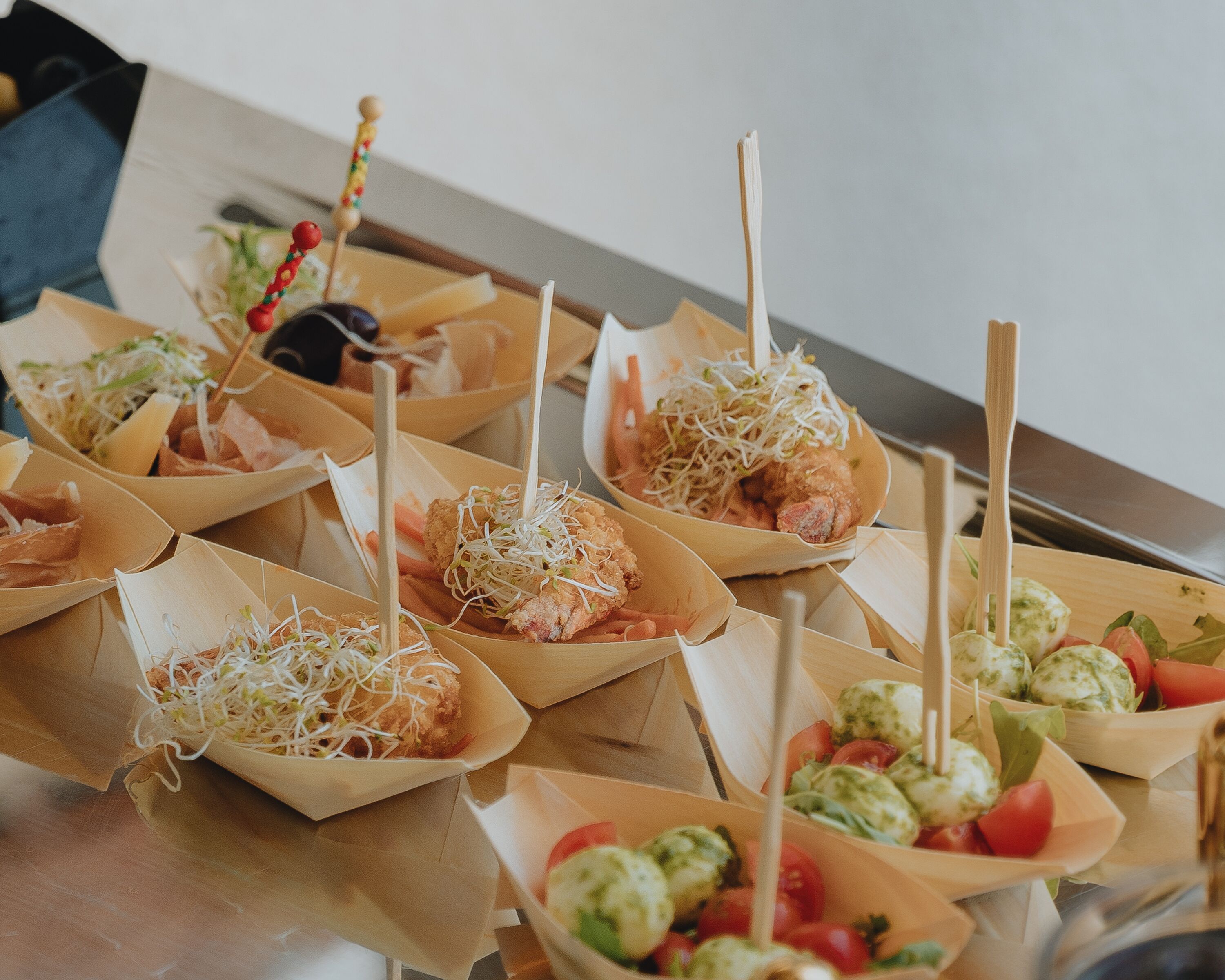 A variety of food items served in paper containers with wooden sticks, including what appears to be fried chicken, sprouts, and vegetables.