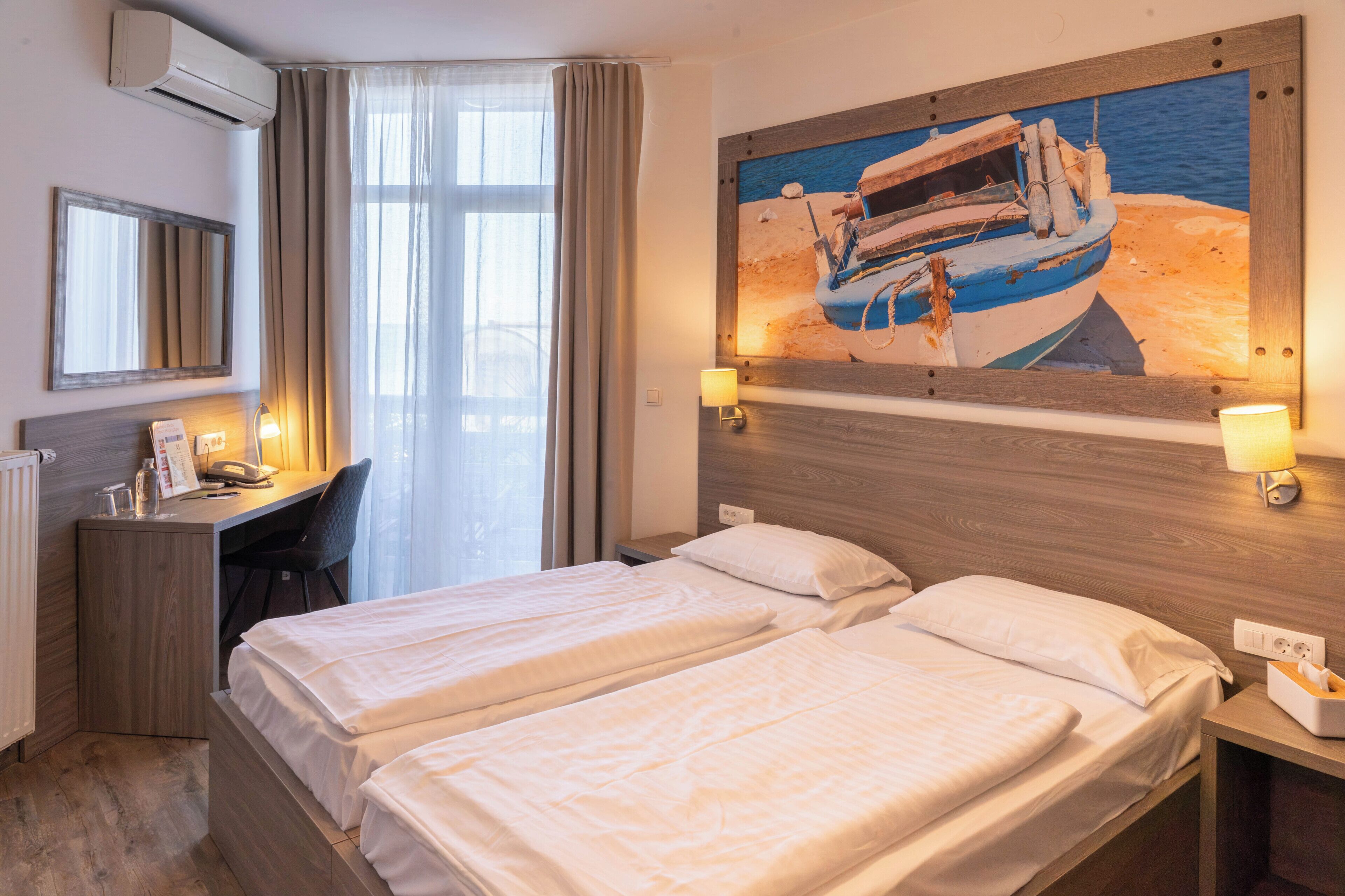 Une chambre d’hôtel avec deux lits, un bureau avec une chaise, une lampe de bureau, un tableau représentant un bateau accroché au mur et une fenêtre avec des rideaux.