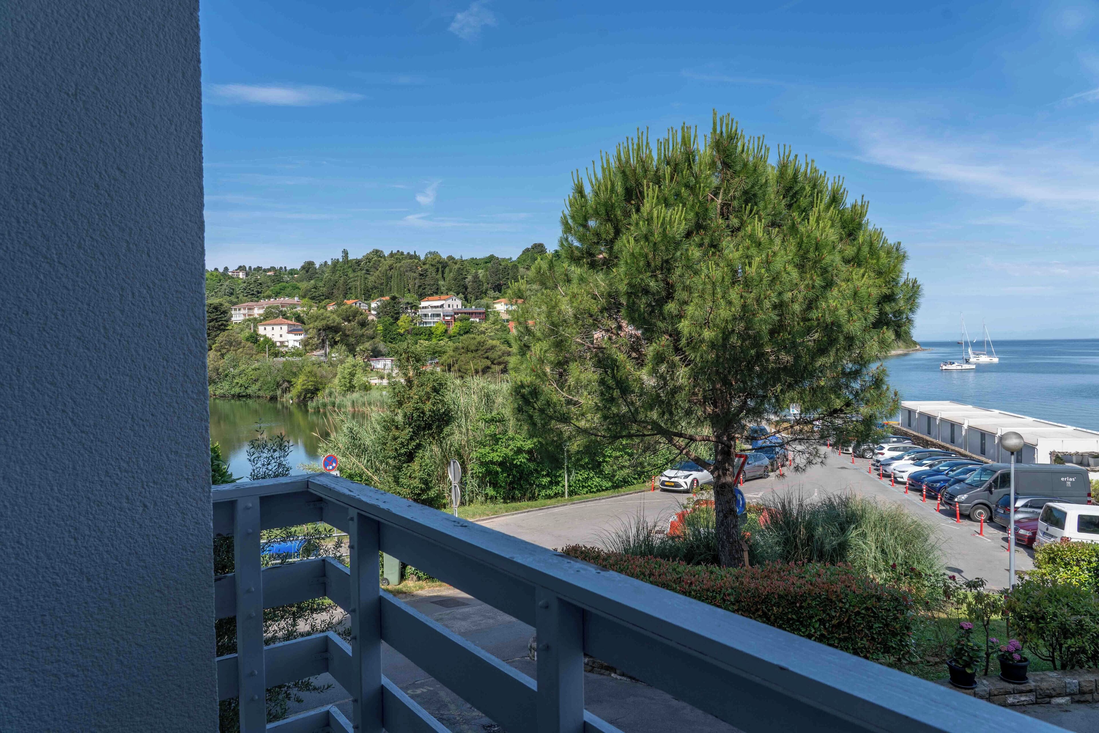 A balcony with a view of a parking area, a tree, and a body of water with boats.