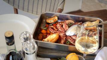 A dining table with a metal tray containing food, a glass of white wine, and a lemon wedge. There are salt and pepper shakers, a bottle of olive oil, and a glass of water on the table.