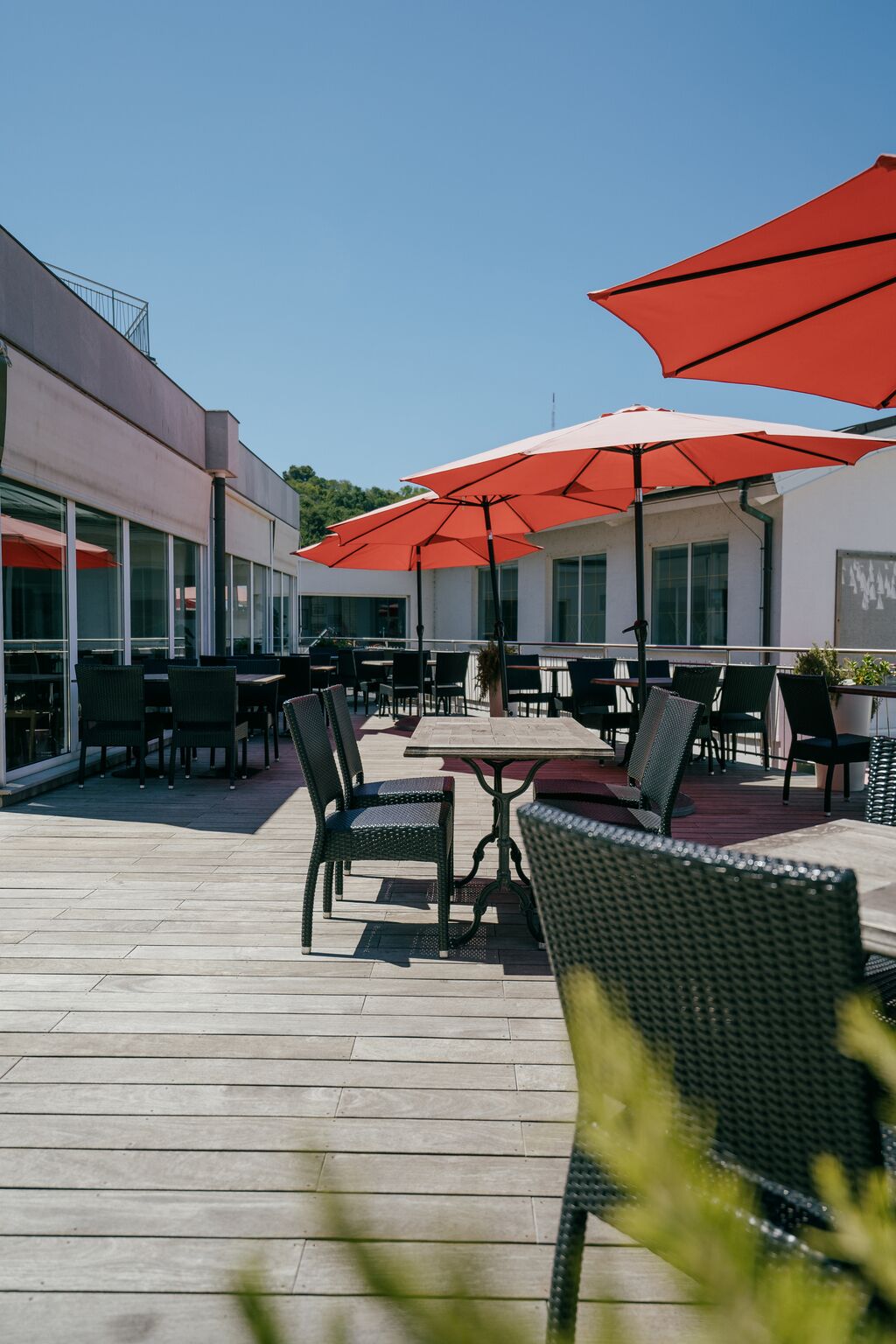 Een terras buiten met tafels en stoelen, rode parasols en een gebouw op de achtergrond.