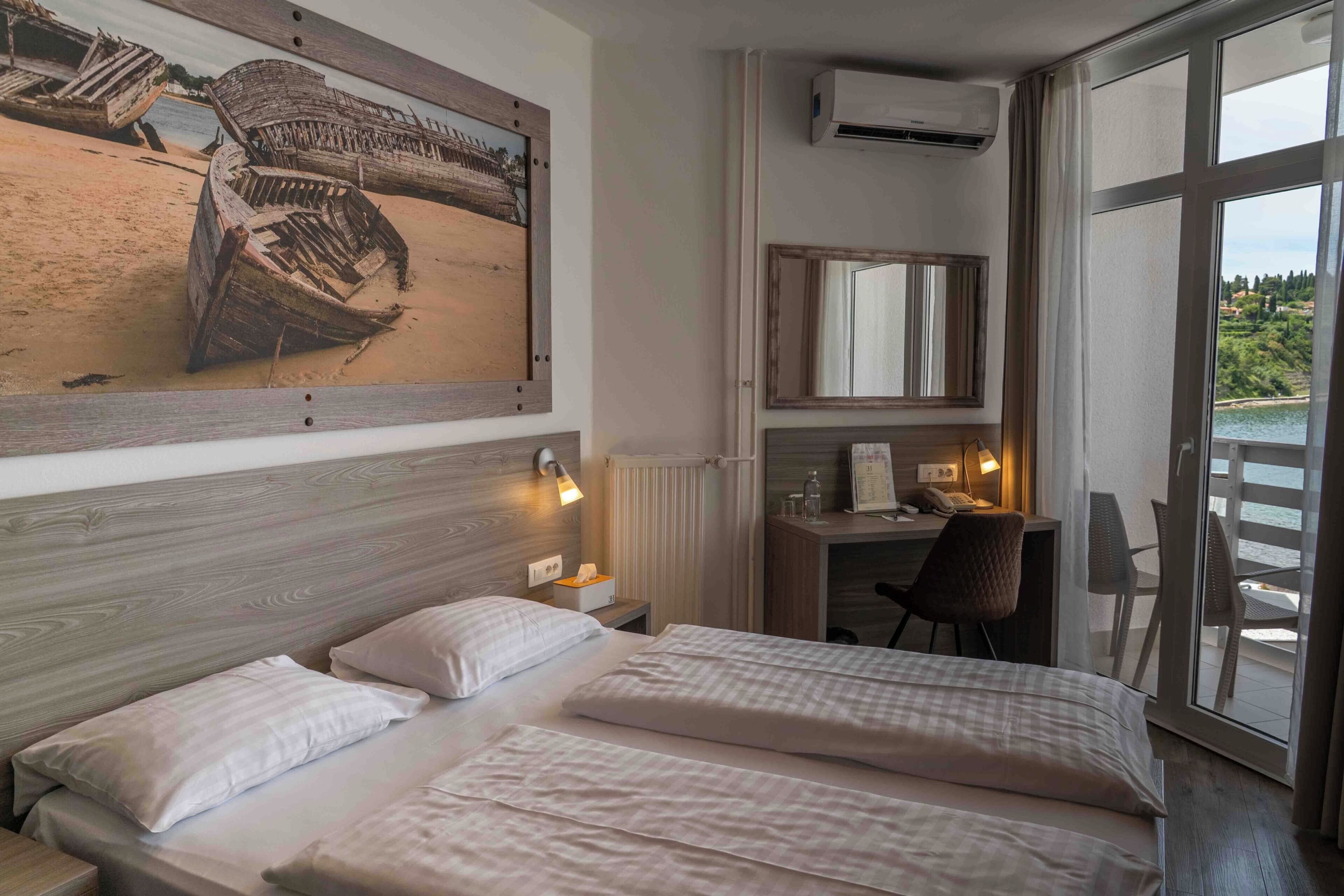 A hotel room with a large bed, a desk with a chair, a mirror, and a view of the sea through large windows.
