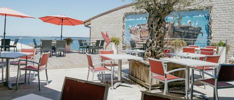 An outdoor patio with tables and chairs, a mural on the wall, and a view of the sea.