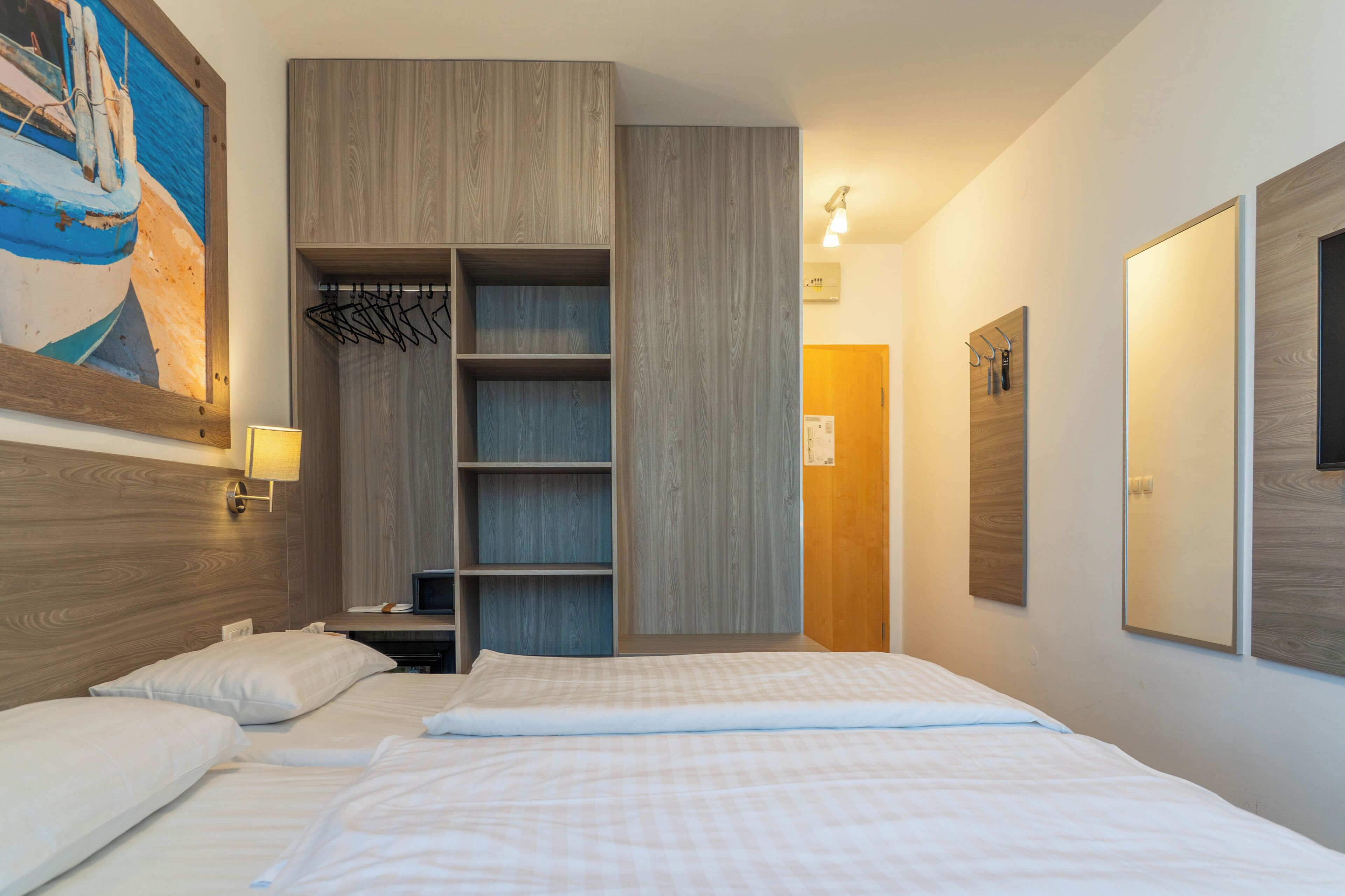 A hotel room with a bed, a wooden wardrobe, a coat rack, and a painting on the wall.