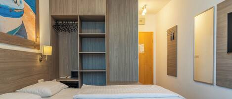 A hotel room with a bed, a wooden wardrobe, a coat rack, and a painting on the wall.