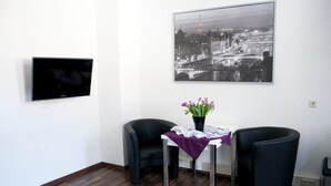 A modern room with a flat-screen TV, a framed cityscape photo, a small table with a vase of flowers, and two black chairs. - Hermann's Neue Post (Calw)