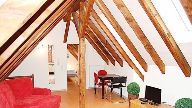 An attic room with exposed wooden beams, a red sofa, a dining table with chairs, and a television on a wooden cabinet. - Hermann's Neue Post (Calw)
