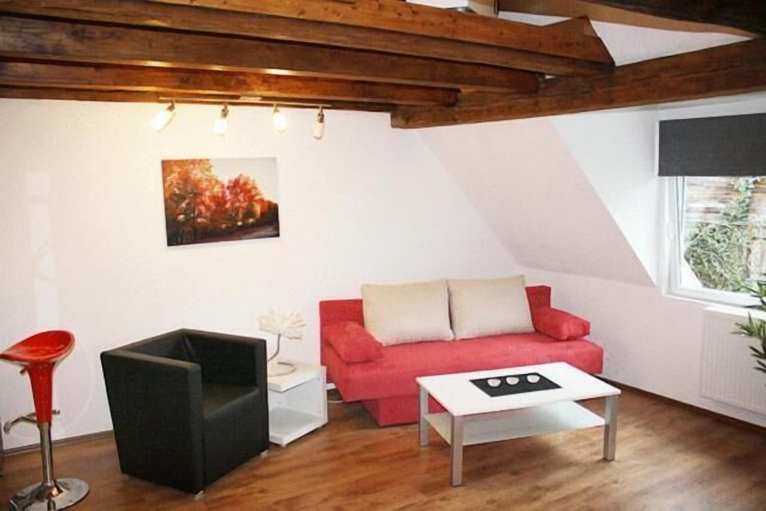 A cozy living room with a red sofa, a black chair, and a white coffee table. There is a painting on the wall and a window with a view of greenery.