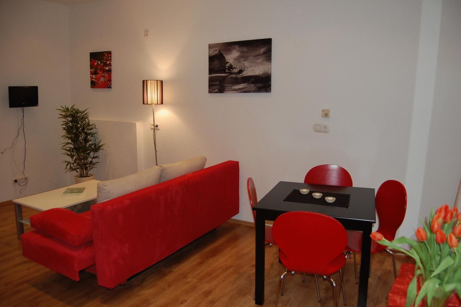 A modern living room with a red sofa, a dining table with red chairs, a lamp, and a painting on the wall.