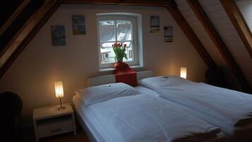 A cozy attic bedroom with a large bed, a nightstand, a window with a view, and a red vase with flowers.