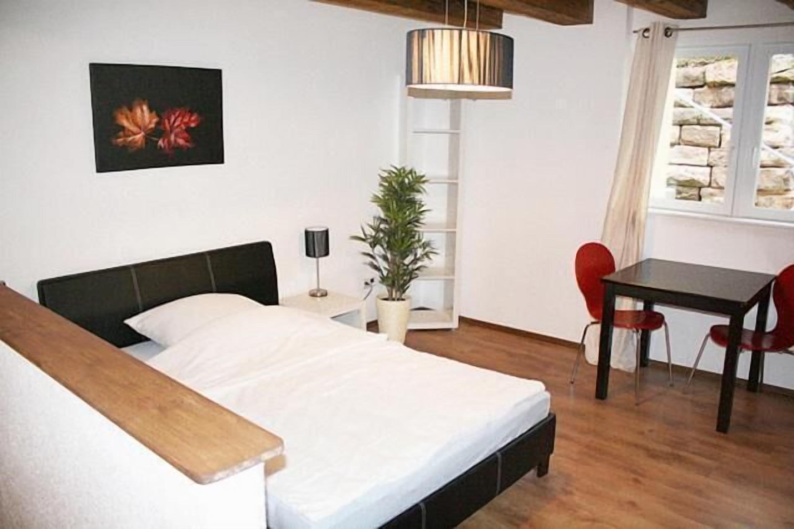 A bedroom with a bed, bedside table, lamp, potted plant, and a dining area with a table and chairs.