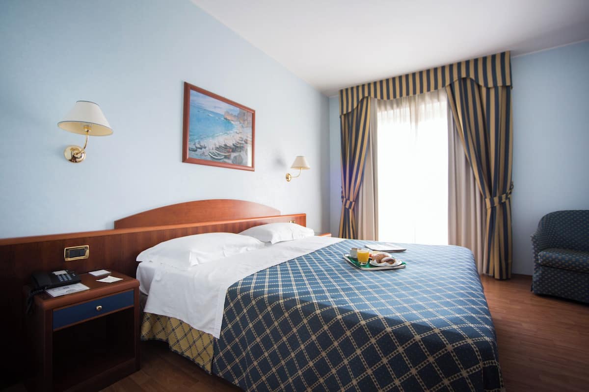 Hotellihuone, jossa on sänky, yöpöytä, jossa on puhelin, lamppu, taulu ja ikkuna, jossa on verhot.