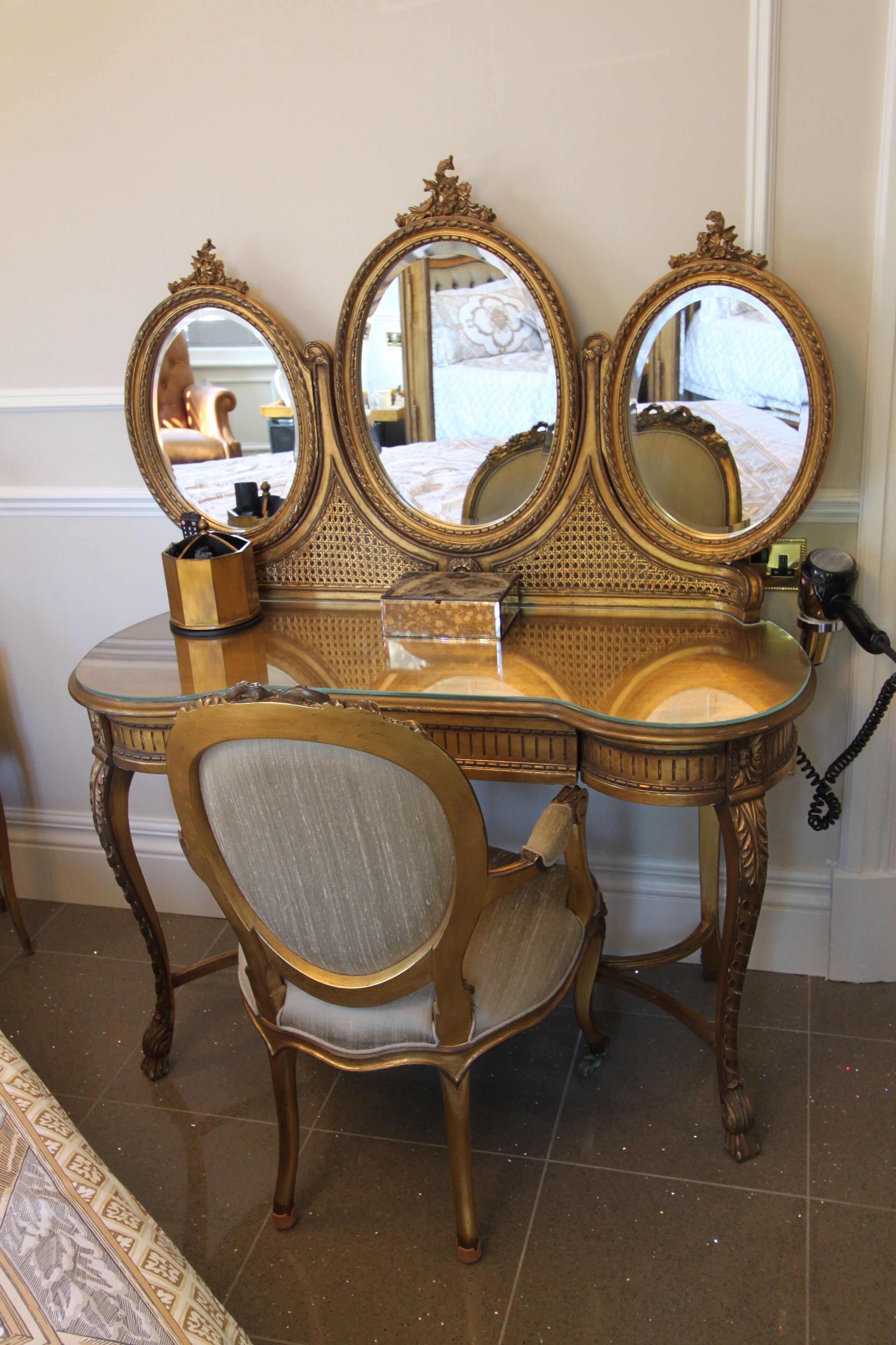 A vanity with three mirrors, a chair, and a small table with a container.