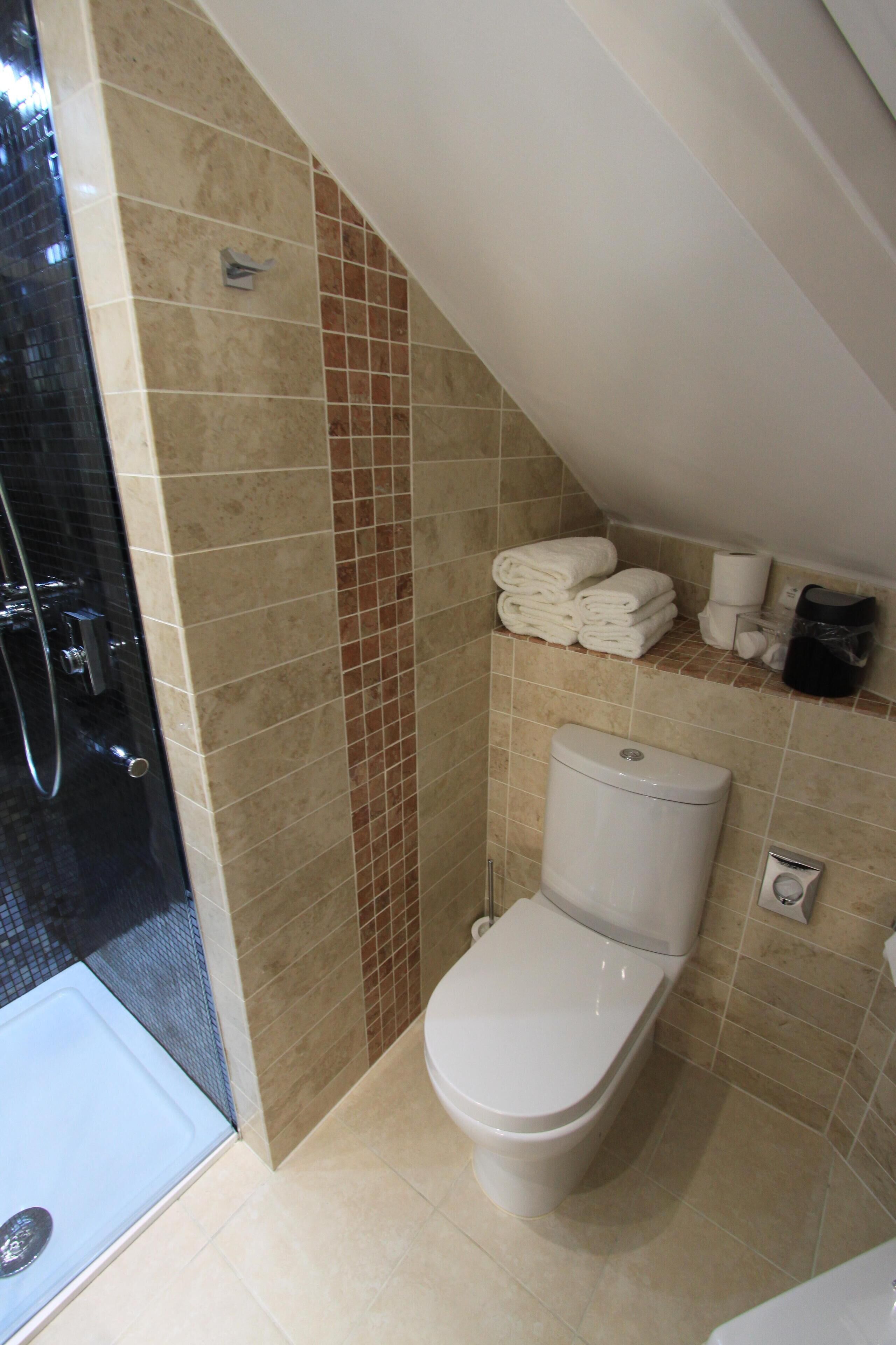 A bathroom with a toilet, a glass shower enclosure, and a tiled wall with a small shelf holding towels and toiletries.