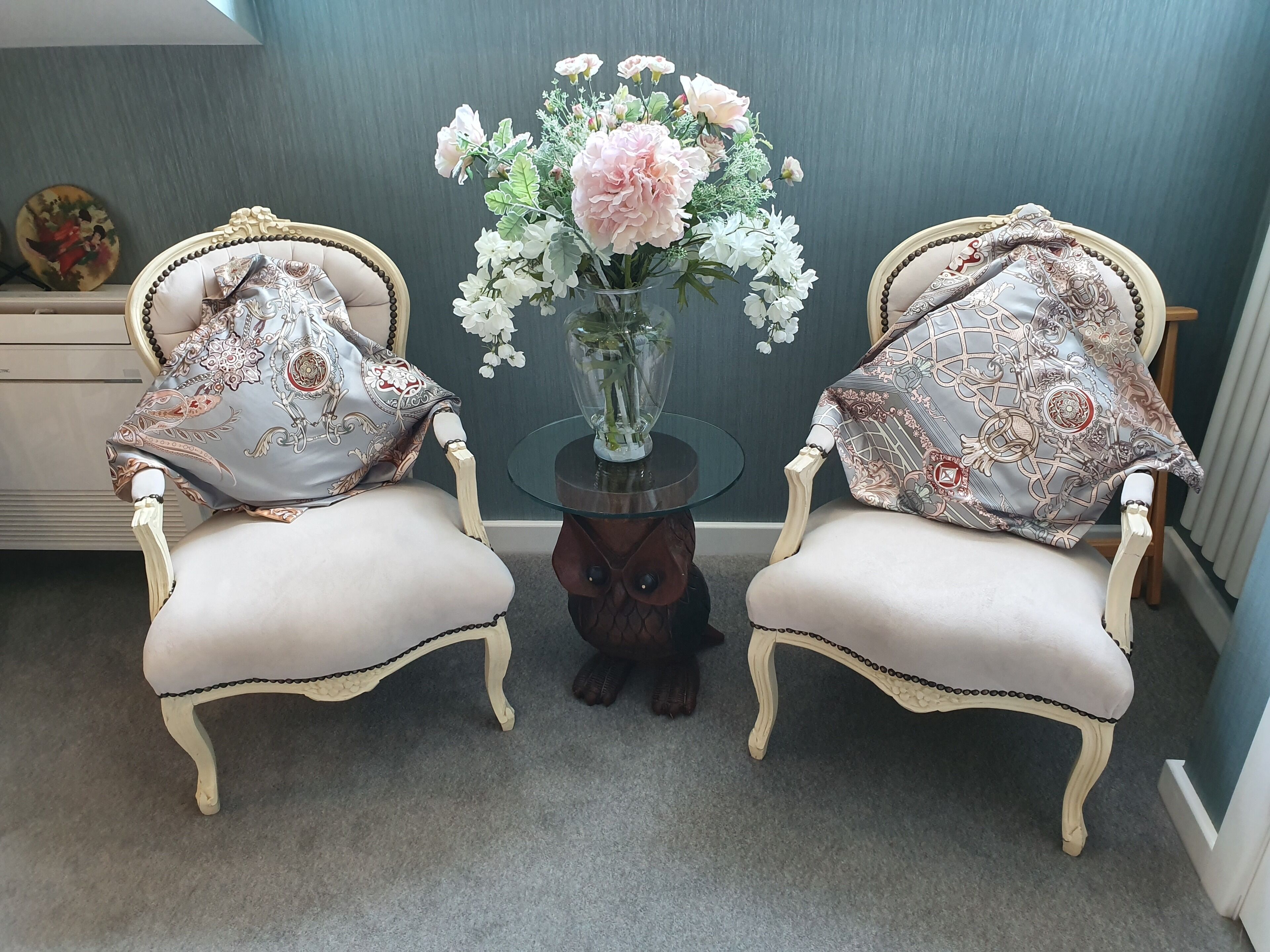 Dos sillones ornamentados con cojines estampados, una mesa de vidrio con un jarrón de flores y un plato decorativo sobre una mesita auxiliar.