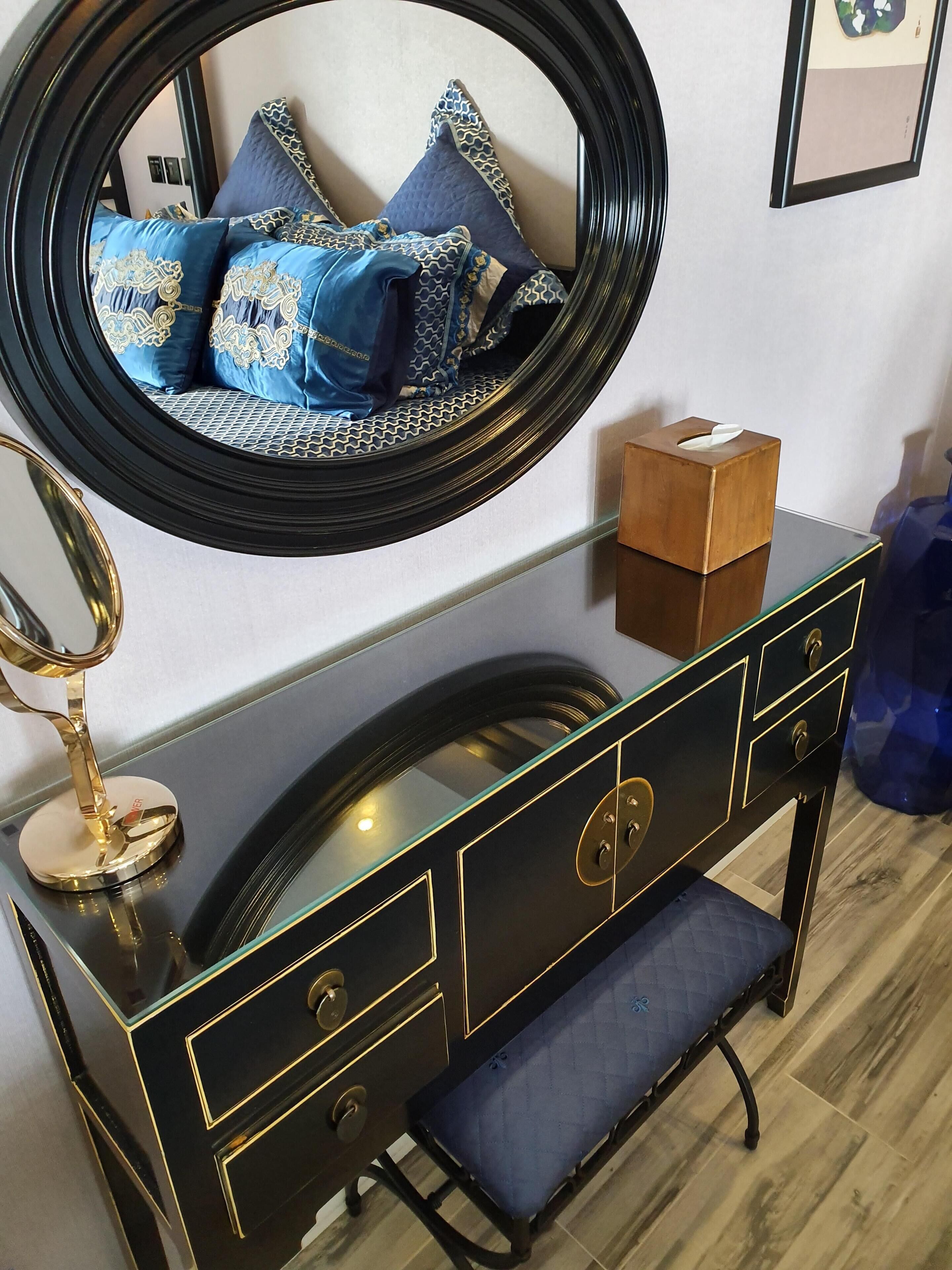 Un tocador con un espejo redondo, una caja de pañuelos de madera y un banco acolchado azul.