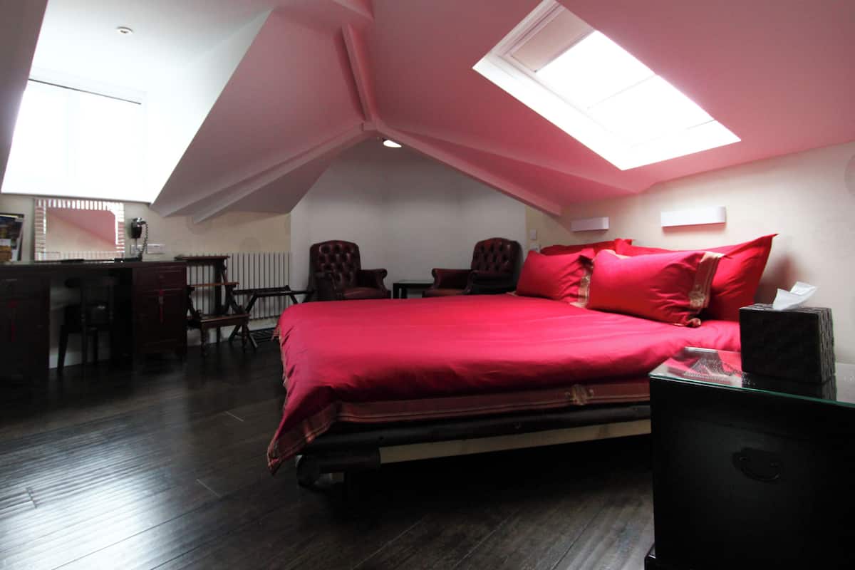 A bedroom with a sloped ceiling, a bed with red bedding, a desk with a chair, and a radiator.