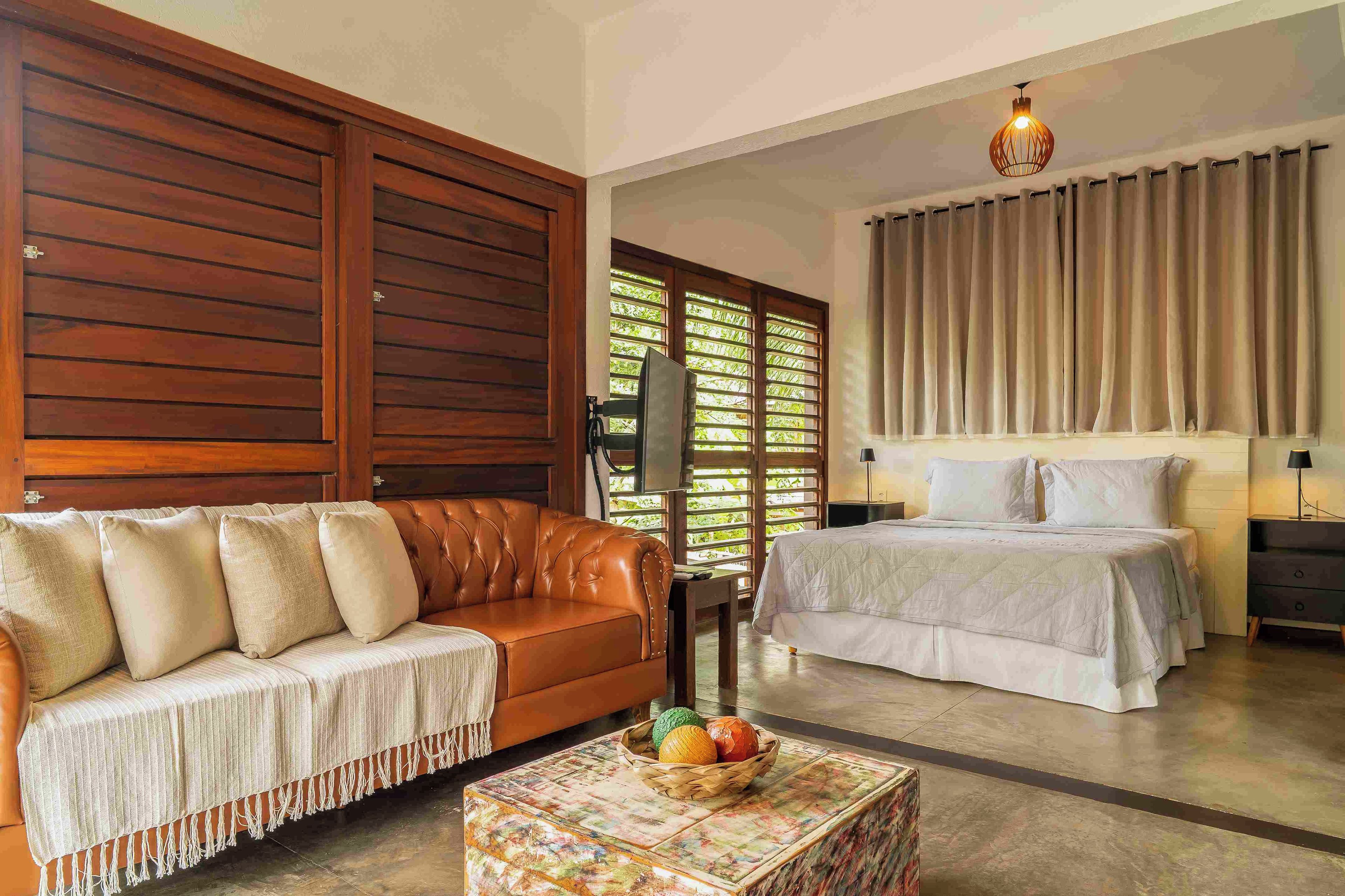 Quarto com sofá de couro marrom, cama com colcha branca, porta de madeira e janela com cortinas.