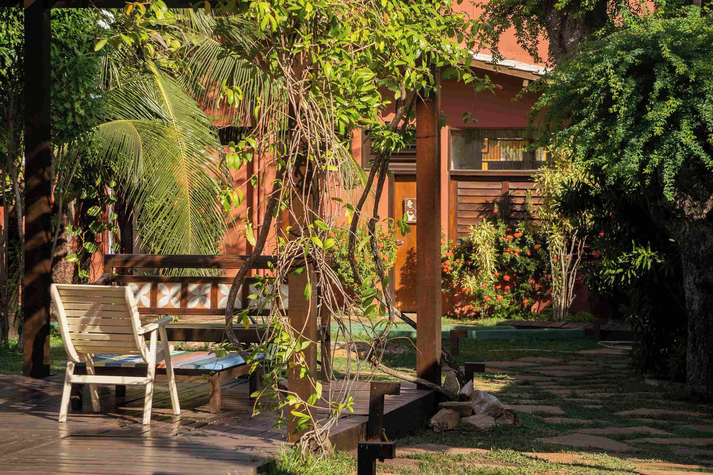 Una terraza de madera con un banco y una mesa, rodeada de abundante vegetación y un edificio con fachada roja.