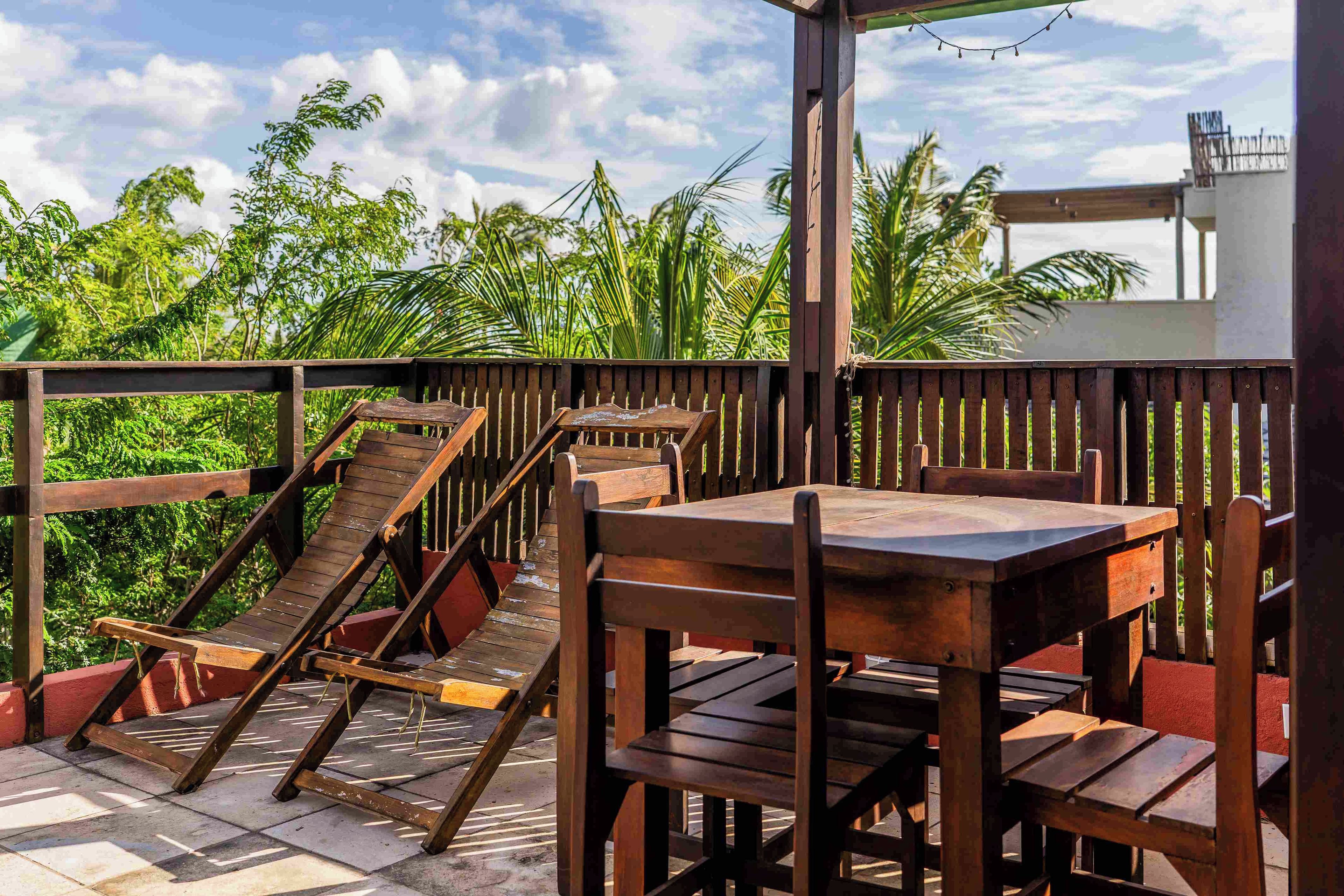 A wooden deck with tables and chairs, surrounded by lush greenery and a clear sky.