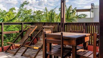A wooden deck with tables and chairs, surrounded by lush greenery and a clear sky.