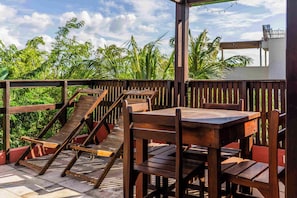 A wooden deck with tables and chairs, surrounded by lush greenery and a clear sky. - Pousada Naquela (Jijoca de Jericoacoara)