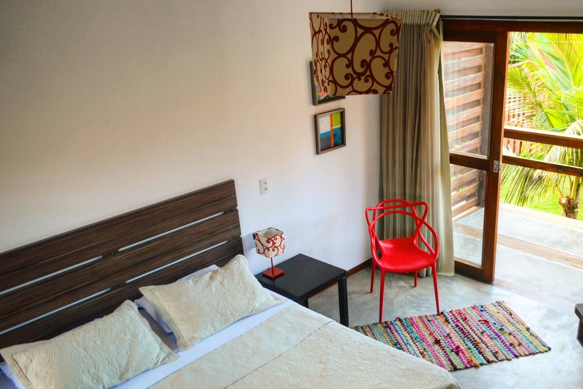 Une chambre à coucher avec un lit, un fauteuil rouge, une petite table avec une lampe, et une vue sur la verdure par la fenêtre.