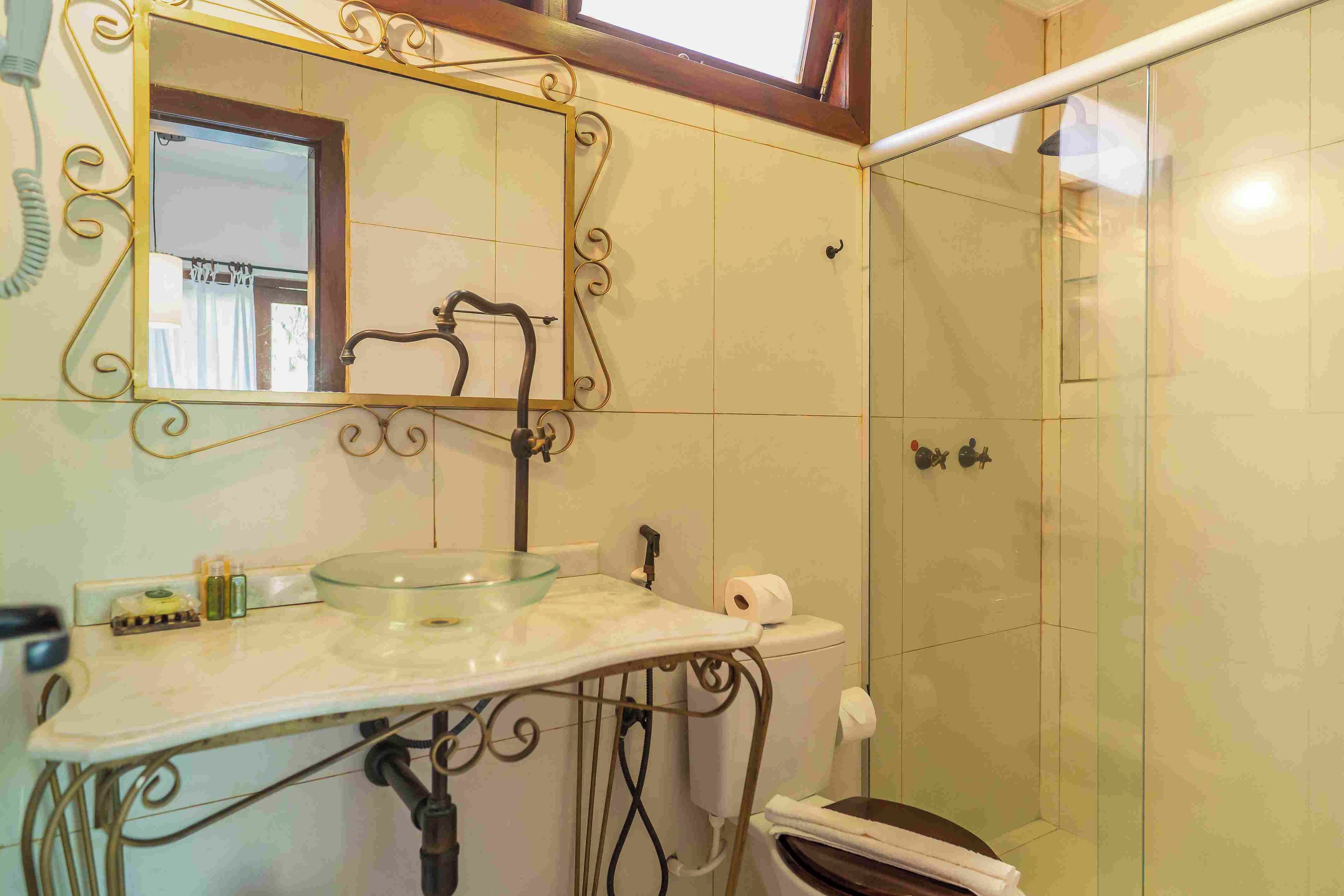 Un baño con lavabo de vidrio, un espejo con detalles de metalurgia ornamentada y zona de ducha.