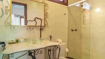 Un baño con lavabo de vidrio, un espejo con detalles de metalurgia ornamentada y zona de ducha.