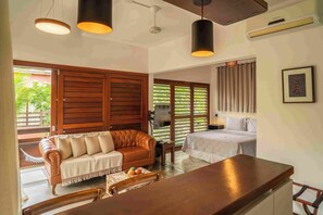 Habitación con sofá de madera, una mesa de comedor y una cama. Se ven ventanas con cortinas y vistas al verdor exterior.