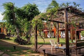 A wooden deck with outdoor seating, surrounded by lush greenery and a building with multiple balconies. - Pousada Naquela (Jijoca de Jericoacoara)