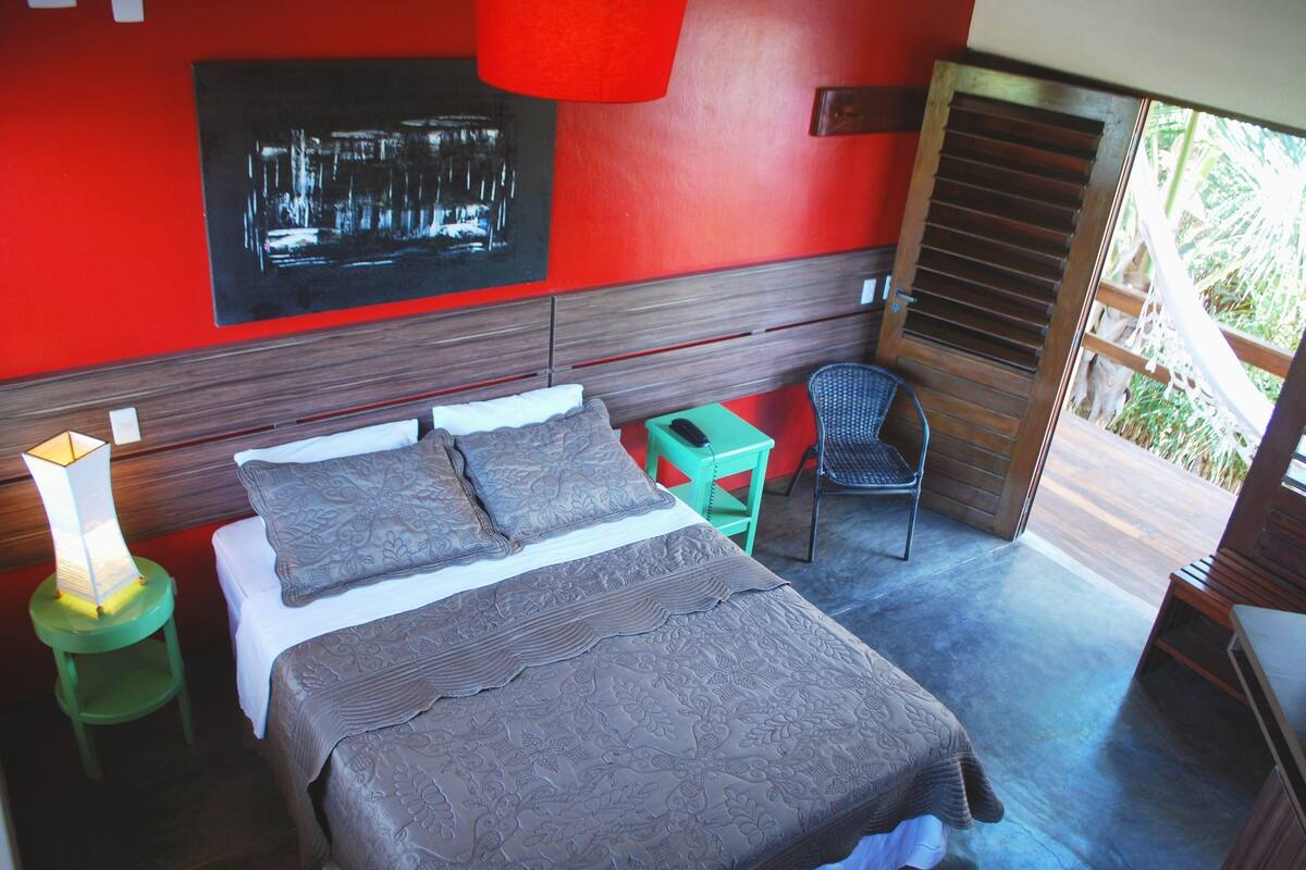 Une chambre à coucher comprenant un lit, une chaise, une table de chevet et un mur rouge orné d’une œuvre d’art noire.