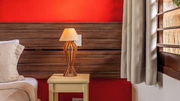 A hotel room with a red wall, a wooden headboard, a bedside table with a lamp, a bed with a white cover, and a window with blinds.