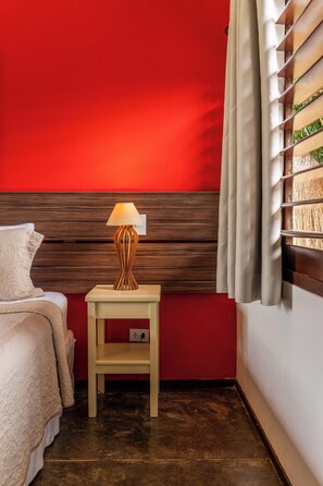 Habitación de hotel con pared roja, cabecera de madera, mesita de noche con lámpara, cama con cubrecama blanco y ventana con persianas.