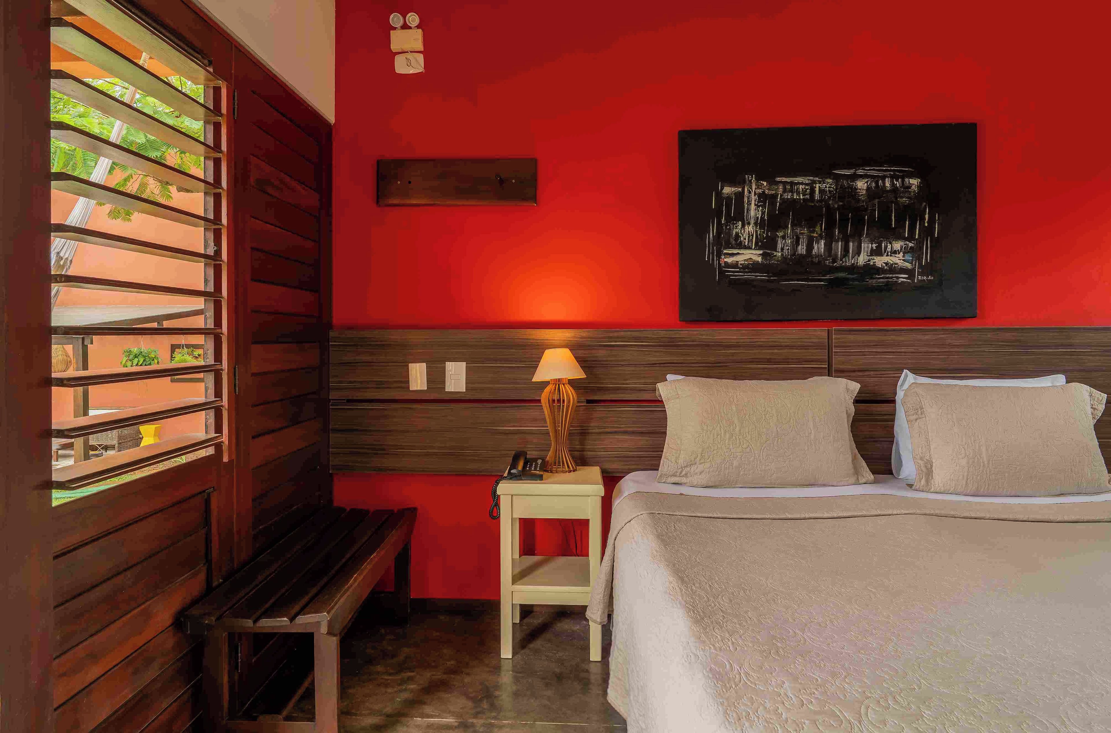 A hotel room with a bed, bedside table, lamp, and a window with a view of greenery.